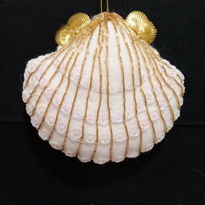 May include: A white seashell ornament with gold beaded accents and gold trim. The ornament is decorated with small, clear sequins and has a gold string for hanging. The seashell is a decorative item.