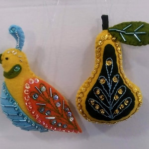 May include: Two handmade felt ornaments: a yellow bird with orange and blue accents, and a yellow pear with a black center. Both are embellished with beads and sequins. These festive decorations are perfect for adding a touch of whimsy to any holiday display.