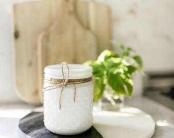 Puede incluir: Un frasco de vidrio blanco con una textura de puntos en relieve en la superficie exterior. El frasco presenta un cuello estrecho envuelto con un cordel de yute marrón natural atado en un pequeño lazo en la parte frontal. El vidrio tiene un acabado opaco.