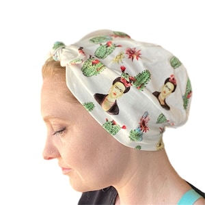May include: A white turban with a print of Frida Kahlo and cacti. The turban is tied in a knot on top of the head.