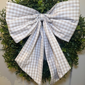 May include: A circular green boxwood wreath adorned with a large, decorative bow featuring a beige and white gingham check pattern. The bow is centered on the foliage, with two wide loops and two long, hanging tails that drape over the greenery.