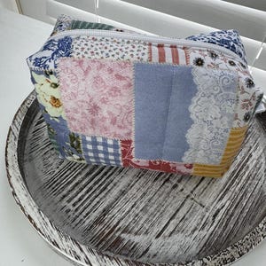 May include: A patchwork cosmetic bag with a zipper closure. The bag features a variety of fabric patterns, including floral, gingham, and lace, in shades of blue, pink, and yellow. The bag is sitting on a round, distressed wooden tray.