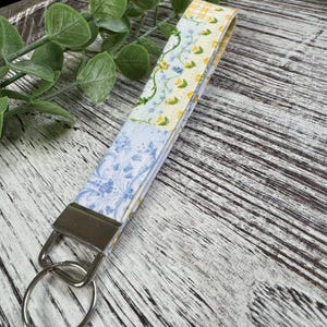 May include: A fabric keychain wristlet with a floral pattern in pastel yellow, blue, and white. The wristlet has a silver-colored metal clasp and key ring. The design features a mix of floral and patterned fabric sections.