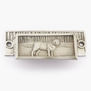 May include: A silver drawer pull with a wolf design. The wolf is standing in a forest setting. The pull has a rectangular shape with rounded corners.