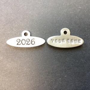 May include: Two oval-shaped silver-toned metal charms with a small circular loop at the top. The left charm is stamped with the number 2026, while the right charm is stamped with the text YOUR NAME in capital letters with a brushed finish.
