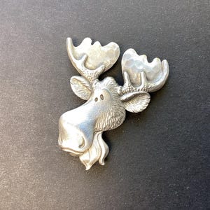 May include: A silver-toned metal brooch shaped like the head of a moose. The design features a textured finish on the antlers and a smooth, polished surface on the face, ears, and neck area, with two small circular indentations for the eyes.