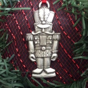 May include: A silver-coloured nutcracker ornament with a red ribbon. The nutcracker is wearing a hat with a feather and has a moustache.