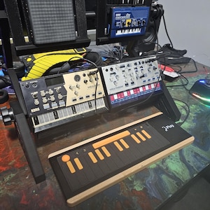 May include: A black and white keyboard with orange keys, a black and white synthesizer with the text "Korg Volca Drum" and a black and white synthesizer with the text "Volca Modular" on a wooden table.