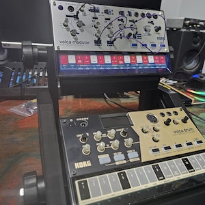 May include: Two black Korg Volca synthesizers, the Volca Modular and the Volca Drum, are stacked on a black stand. The Volca Modular has a silver faceplate with black knobs and buttons. The Volca Drum has a gold faceplate with black knobs and buttons. Both synthesizers have a row of keys.
