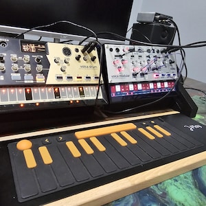 May include: Electronic music production setup featuring Korg Volca Drum and Volca Modular synthesizers, along with a black and orange keyboard controller. The setup includes various cables and a small speaker, all arranged on a patterned surface.