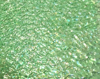 May include: A sheet of textured glass with a light green tint, featuring a rippled, uneven surface that creates a wavy, water-like appearance with numerous small, rounded indentations and reflective highlights across the entire surface.