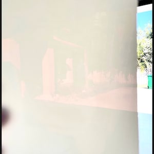 May include: A soft, off-white, translucent material, possibly a sheet or panel, dominates the image. The material is slightly blurred, with hints of a structure and outdoor elements visible through it. The edges are framed in black.