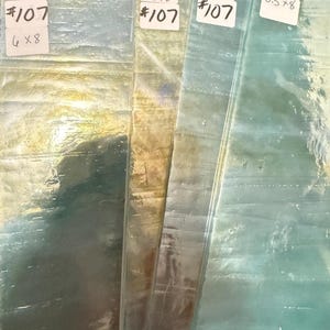 以下が含まれることがあります： 青と緑の異なる色合いの虹色のガラスが3枚。ガラスは質感があり、かすかな輝きを放っています。各ピースには「#107」と「6 x 8」、「5.5 x 8」、「5.5 x 8」の寸法が記されています。