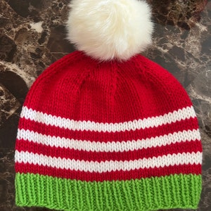 May include: Red and white striped knit beanie with a green brim and a white faux fur pom pom.
