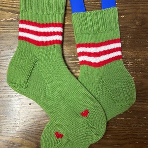 May include: A pair of green knitted socks with red and white stripes around the ankle. The socks have red hearts on the toes.