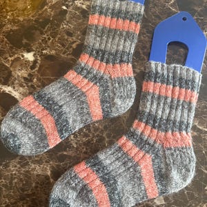 May include: A pair of grey and coral striped socks. The socks are made of a thick, textured material and are displayed on blue hangers. The socks have a ribbed design on the cuff and foot.
