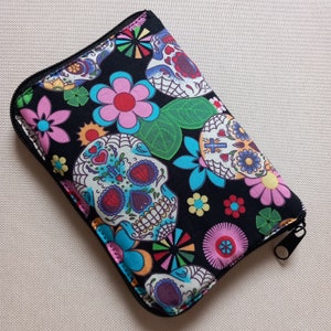 May include: A black zippered wallet with a vibrant Day of the Dead sugar skull and floral design. The pattern includes pink, blue, and green flowers, and a black zipper. The wallet is rectangular in shape.