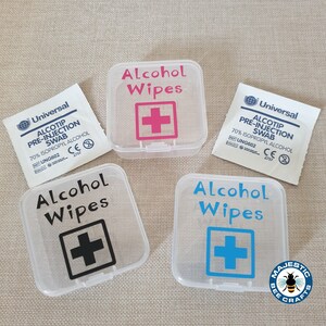 K&ouml;nnte beinhalten: Vier durchsichtige Plastikbeh&auml;lter mit dem Text "Alcohol Wipes" und einem Pluszeichen in verschiedenen Farben. Die Beh&auml;lter sind mit dem Text "Alcohol Wipes" und einem Pluszeichen in Schwarz, Pink, Blau und Schwarz beschriftet. Die Beh&auml;lter befinden sich auf einer braunen Oberfl&auml;che.