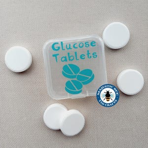 May include: A clear plastic container with a white lid labelled "Glucose Tablets". The container has three blue tablets inside. There are five white tablets outside the container on a beige surface. The container has a sticker on the bottom that says "Majestic Bee T1D Crafts".