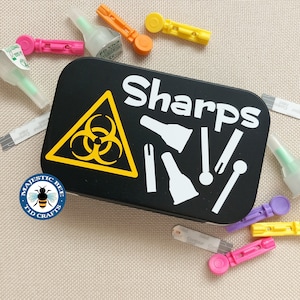 Peut inclure: Un récipient à aiguilles noir avec un symbole de risque biologique jaune et le mot "Sharps" en lettres blanches. Le récipient est entouré de divers fournitures médicales, notamment des lancettes, des dispositifs de lancettes et des bandelettes de test.