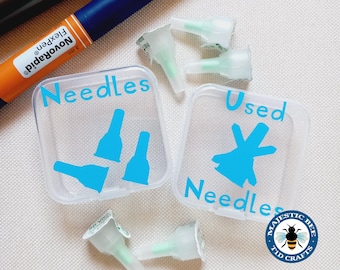 May include: Two clear plastic containers with blue labels that say "Needles" and "Used Needles". The containers are for storing insulin needles. There are several insulin needles scattered around the containers.