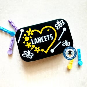 May include: A black rectangular storage tin with the word LANCETS printed in white inside a yellow heart outline, surrounded by white bee graphics and yellow flowers. Several small, colourful plastic lancets are scattered around the tin.