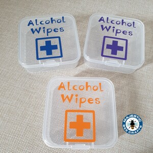 K&ouml;nnte beinhalten: Drei transparente Plastikbeh&auml;lter mit dem Text "Alcohol Wipes" und einem roten Kreuz darauf gedruckt. Die Beh&auml;lter sind mit einem blauen, violetten und orangefarbenen Kreuz gekennzeichnet.