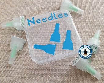 May include: A clear plastic container with the word "Needles" printed on the lid. The container holds three blue plastic needle caps. The container is sitting on a beige fabric surface. There are four clear plastic needle caps with green tips scattered around the container.