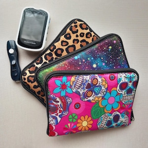 May include: Three patterned zippered pouches: a leopard print, a galaxy design, and a pink sugar skull print with flowers. A blood glucose meter and a lancet device are also visible.