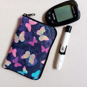May include: A black blood glucose meter, a white and black lancing device, and a zippered pouch with a butterfly pattern. The pouch is navy blue with pink, yellow, and blue butterflies.