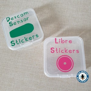 May include: Two clear plastic square containers with lids. The container on the left has a green sensor sticker with the text "Dexcom Sensor Stickers". The container on the right has a pink circle sticker with the text "Libre Stickers".