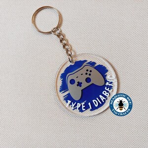 Peut inclure: Porte-clés rond en acrylique transparent avec un motif de manette de jeu argentée sur fond bleu. Le texte "TYPE 1 DIABETIC" est en blanc. Il est muni d'un porte-clés et d'une chaîne argentés. Le porte-clés a également un logo qui dit "MAJESTIC BEE TID CRAFTS".