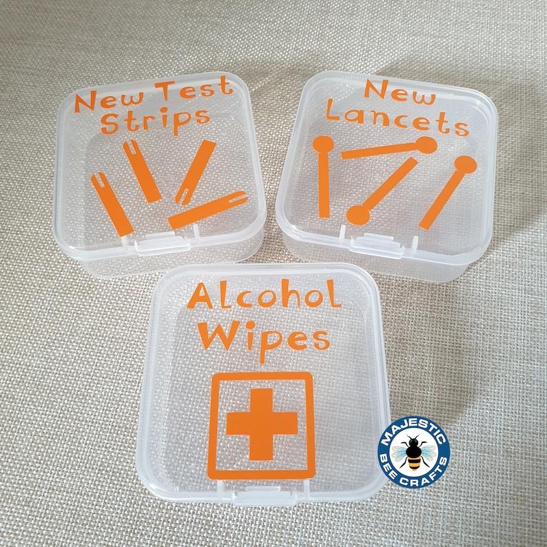 K&ouml;nnte beinhalten: Drei transparente Plastikbeh&auml;lter mit orangenen Etiketten. Die Beh&auml;lter sind mit "Neue Teststreifen", "Neue Lanzetten" und "Alkoholtupfer" beschriftet. Der Beh&auml;lter mit der Aufschrift "Alkoholtupfer" hat auch ein rotes Kreuzsymbol. Die Beh&auml;lter befinden sich auf einer beigefarbenen Oberfl&auml;che.