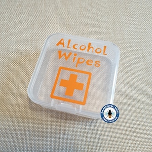 K&ouml;nnte beinhalten: Ein durchsichtiger Plastikbeh&auml;lter mit einem wei&szlig;en Etikett, auf dem "Alcohol Wipes" und ein rotes Kreuzsymbol stehen. Der Beh&auml;lter befindet sich auf einem braunen Stoffhintergrund.
