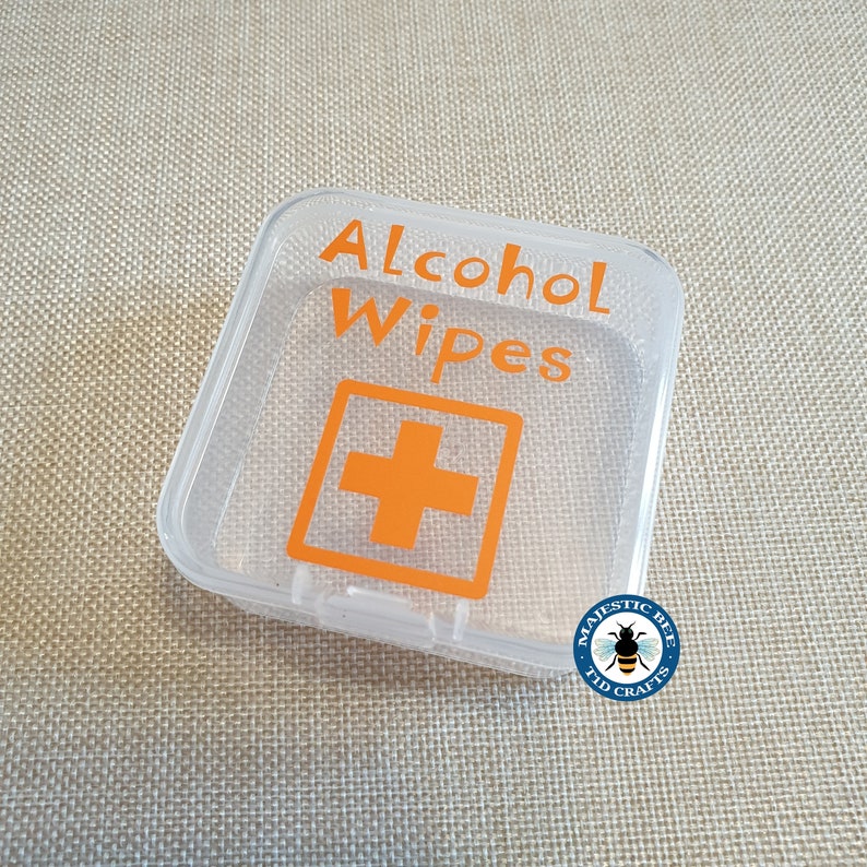K&ouml;nnte beinhalten: Ein durchsichtiger Plastikbeh&auml;lter mit einem wei&szlig;en Etikett, auf dem "Alcohol Wipes" und ein rotes Kreuzsymbol stehen. Der Beh&auml;lter befindet sich auf einem braunen Stoffhintergrund.