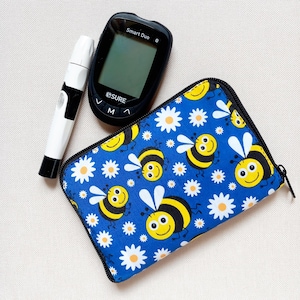 May include: A diabetes testing kit comprising a black and white lancing device, a black Smart Duo blood glucose meter, and a blue zippered case. The case is patterned with yellow bees and white daisies.