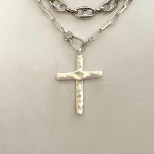 May include: A layered silver chain necklace with a hammered silver cross pendant. The necklace features three different chain styles, including a curb chain, a box chain, and a cable chain. The cross pendant is a simple design with a textured finish.