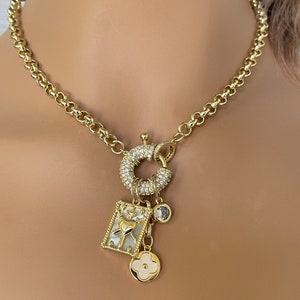 May include: A gold-toned necklace with a chunky chain and a large, sparkling clasp. The necklace features several charms, including a square pendant with a heart design, a round charm, and a four-leaf clover charm. The necklace is displayed against a neutral background.