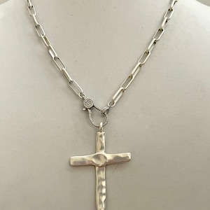 May include: A silver chain necklace with a hammered silver cross pendant. The chain has a clasp with small clear stones.