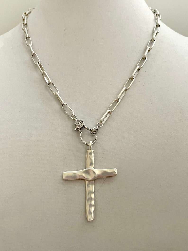 May include: A silver chain necklace with a hammered silver cross pendant. The chain has a clasp with small clear stones.