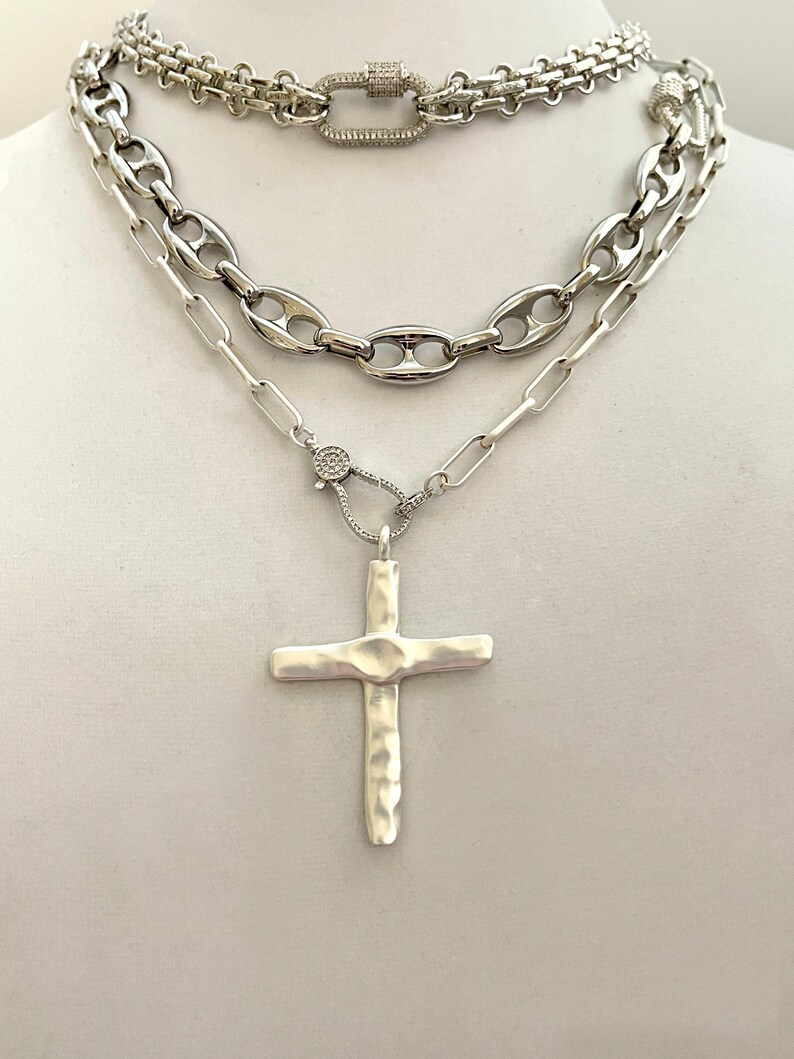 May include: A layered silver chain necklace with a hammered silver cross pendant. The necklace features three different chain styles, including a curb chain, a box chain, and a cable chain. The cross pendant is a simple design with a textured finish.