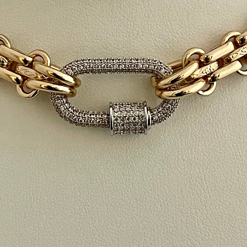 May include: A gold chain necklace with a large oval-shaped clasp set with small clear stones. The clasp is made of two interlocking pieces.