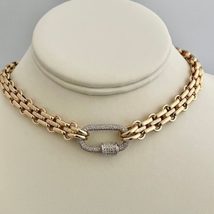 May include: A gold chain necklace with a large, oval-shaped clasp set with small, clear diamonds. The chain is made of multiple rows of linked metal.