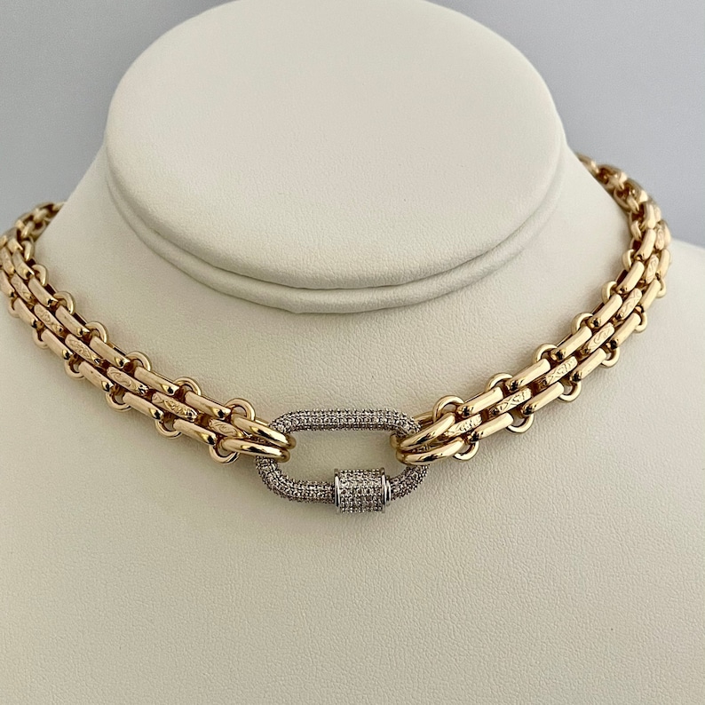 May include: A gold chain necklace with a large, oval-shaped clasp set with small, clear diamonds. The chain is made of multiple rows of linked metal.