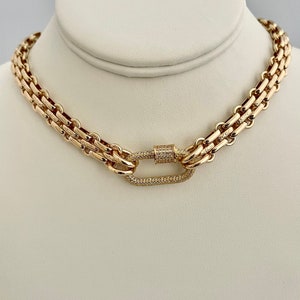 May include: A gold chain necklace with a large, oval-shaped clasp set with small diamonds. The chain is made of two rows of linked squares.