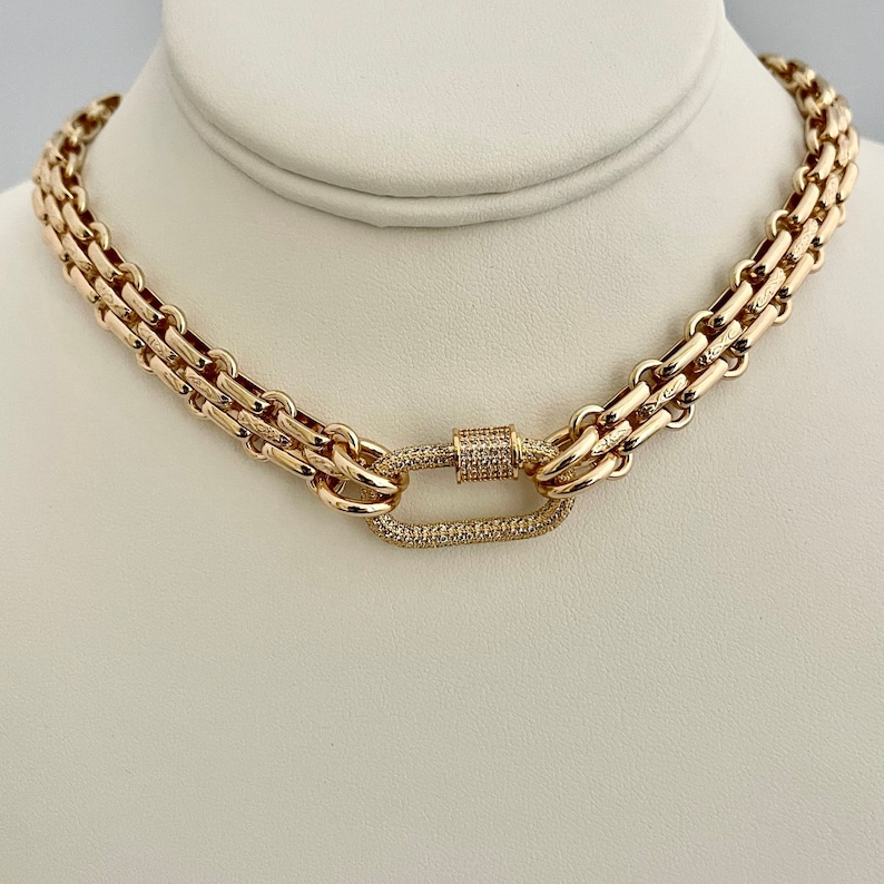 May include: A gold chain necklace with a large, oval-shaped clasp set with small diamonds. The chain is made of two rows of linked squares.