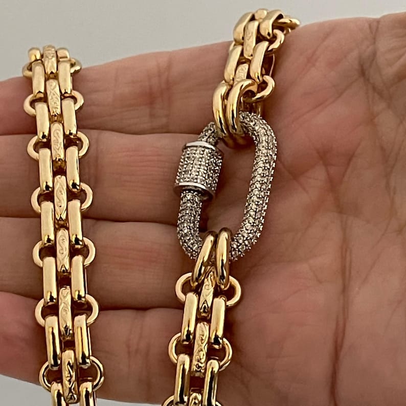 May include: A gold chain necklace with a large, oval-shaped clasp set with clear stones. The chain is made of multiple rows of linked gold pieces.