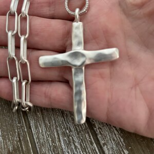 May include: A silver chain necklace with a large, hammered silver cross pendant. The chain is a box chain style.