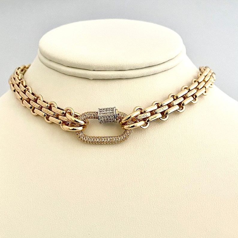 May include: A gold chain necklace with a large, oval-shaped clasp set with small diamonds. The chain is made of two rows of linked gold squares.
