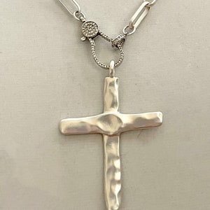 May include: A silver chain necklace with a hammered silver cross pendant and a silver clasp with small clear stones.